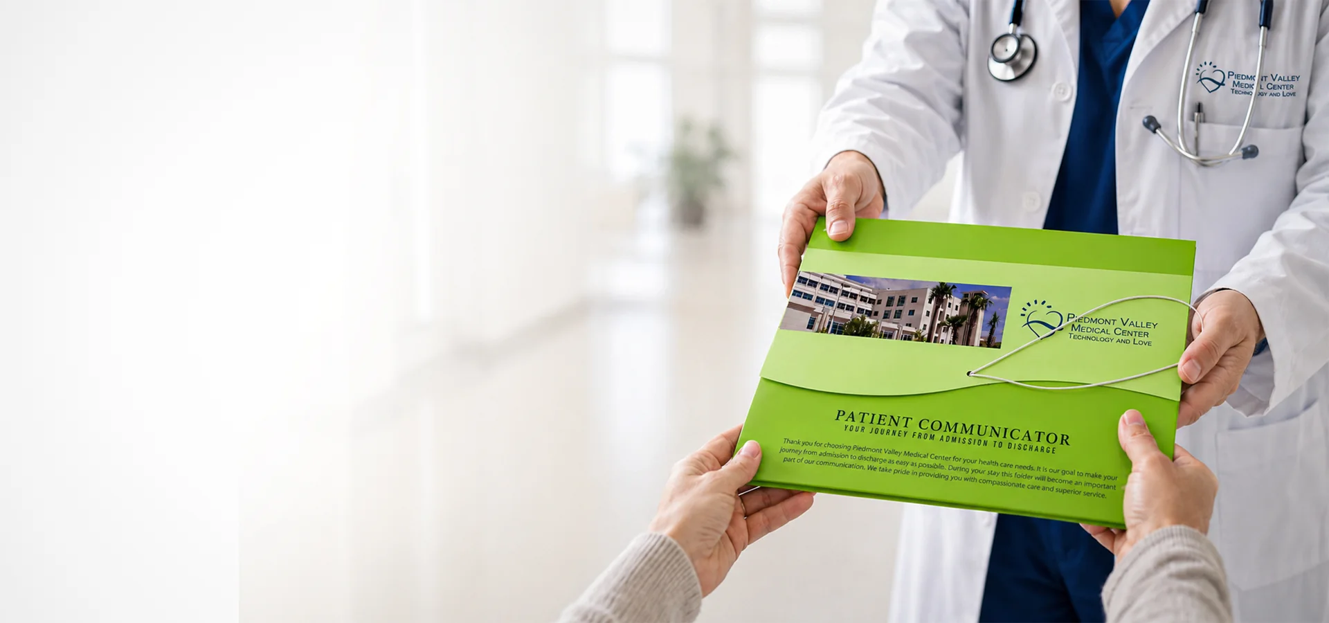 A physician in an open white coat and stethoscope handing a large green Piedmont Valley Medical Center Patient Communicator admit and discharge folder to a patient